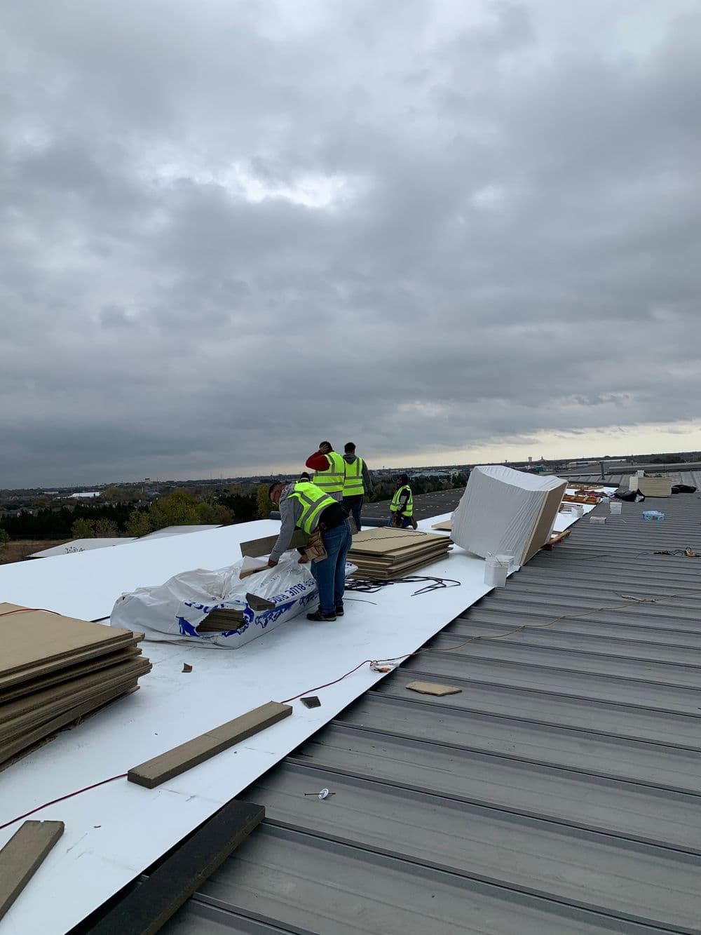 Workers installing roofing materials on a flat roof under overcast skies.