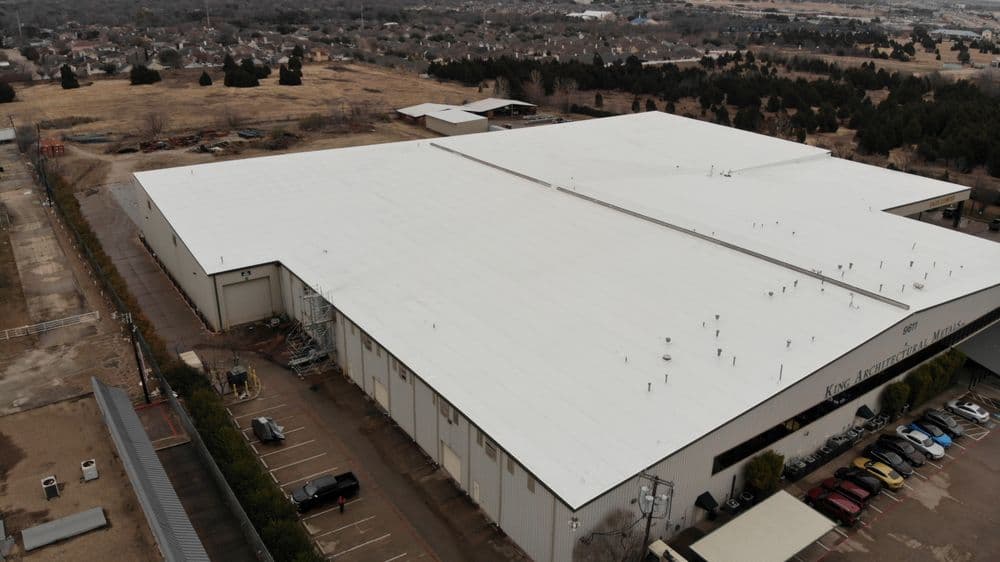 Aerial view of a large industrial building with a white roof and surrounding landscape.