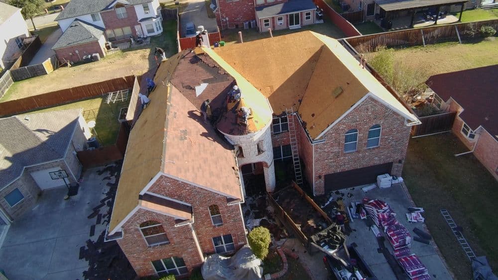 Aerial view of a large brick house undergoing roof replacement with workers and materials visible.