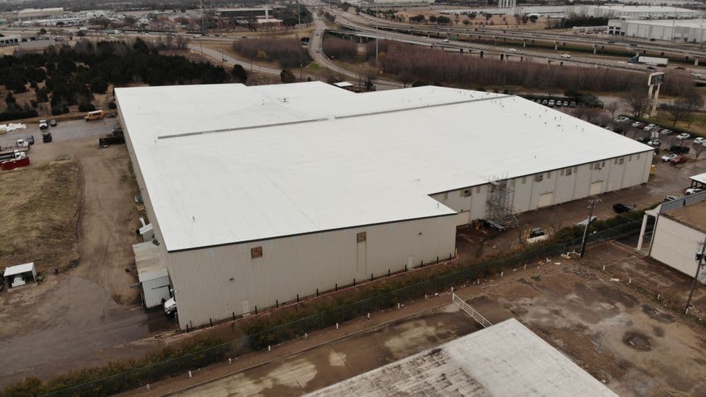 Aerial view of a large industrial building with a white roof and surrounding landscape.