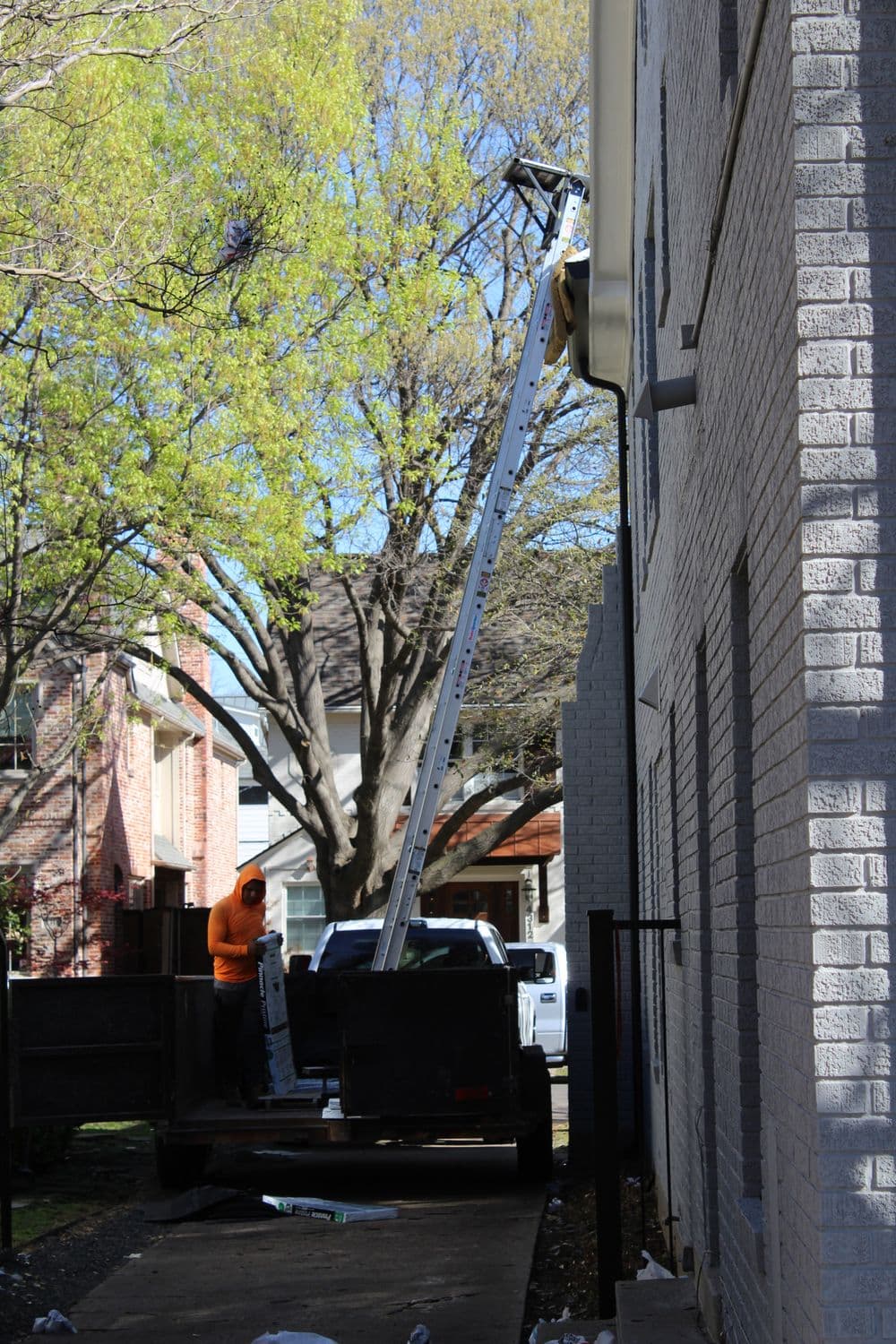 Worker on a ladder cleaning gutters next to a house with trees in the background.