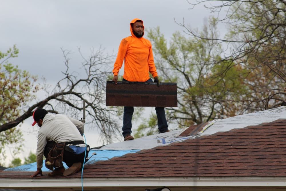 Workers repairing a roof, one holding materials while standing on the roof.