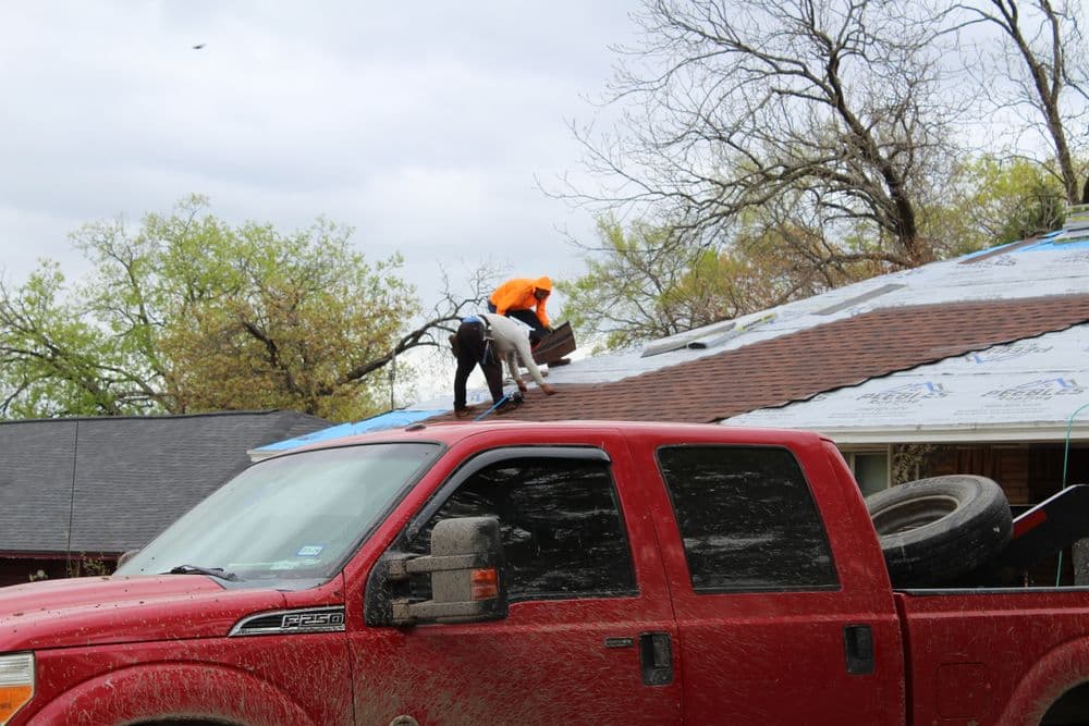Workers installing a roof on a house, with a red truck parked below.
