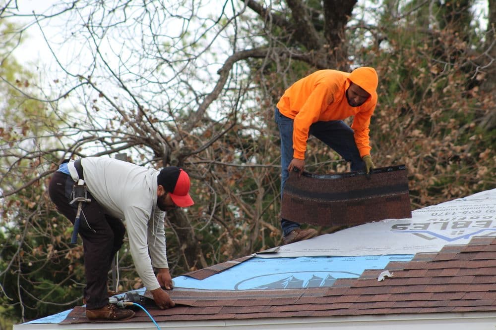 Two workers installing roofing shingles on a house, one standing and one kneeling.