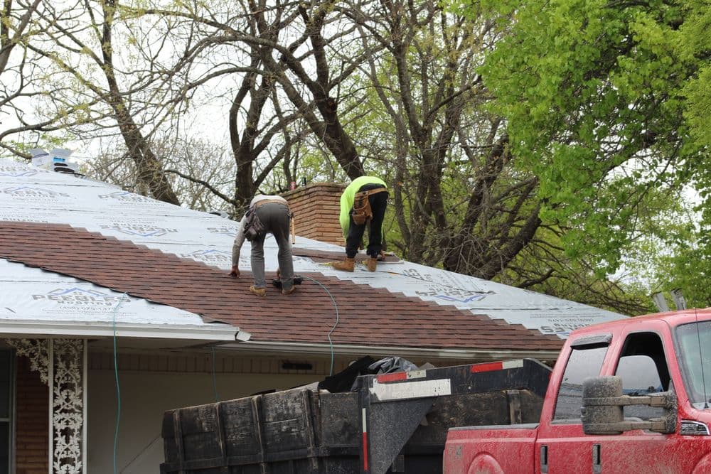 Roofers installing shingles on a residential home, with a red truck and debris trailer nearby.