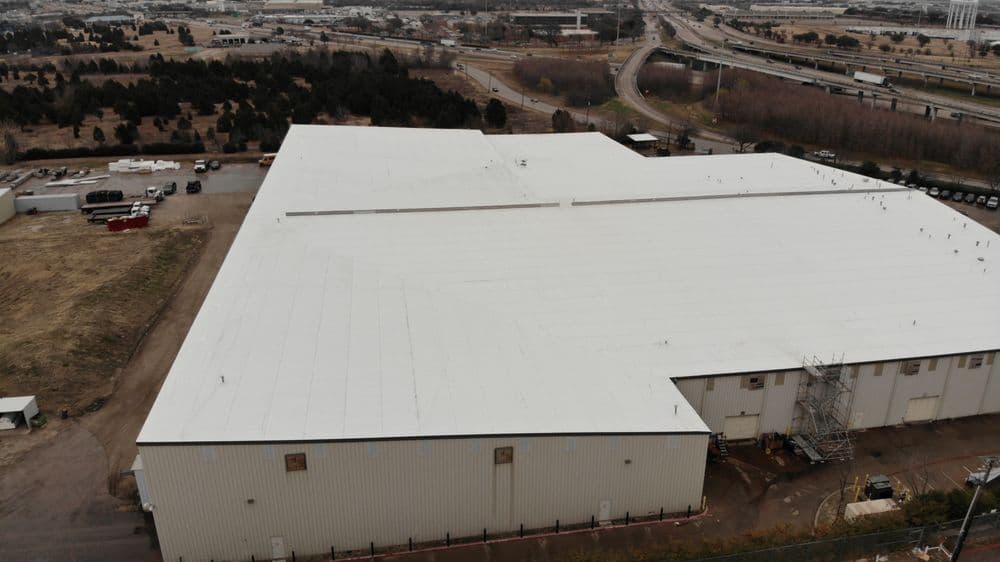 Aerial view of a large industrial warehouse with a white roof and surrounding landscape.