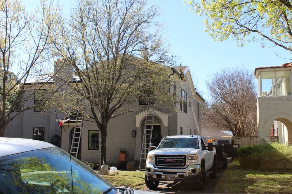 Residential home with ladders, surrounded by trees and parked vehicles on a sunny day.