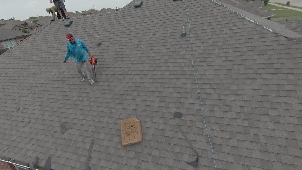 Worker on a roof using a leaf blower, surrounded by shingles and residential houses.
