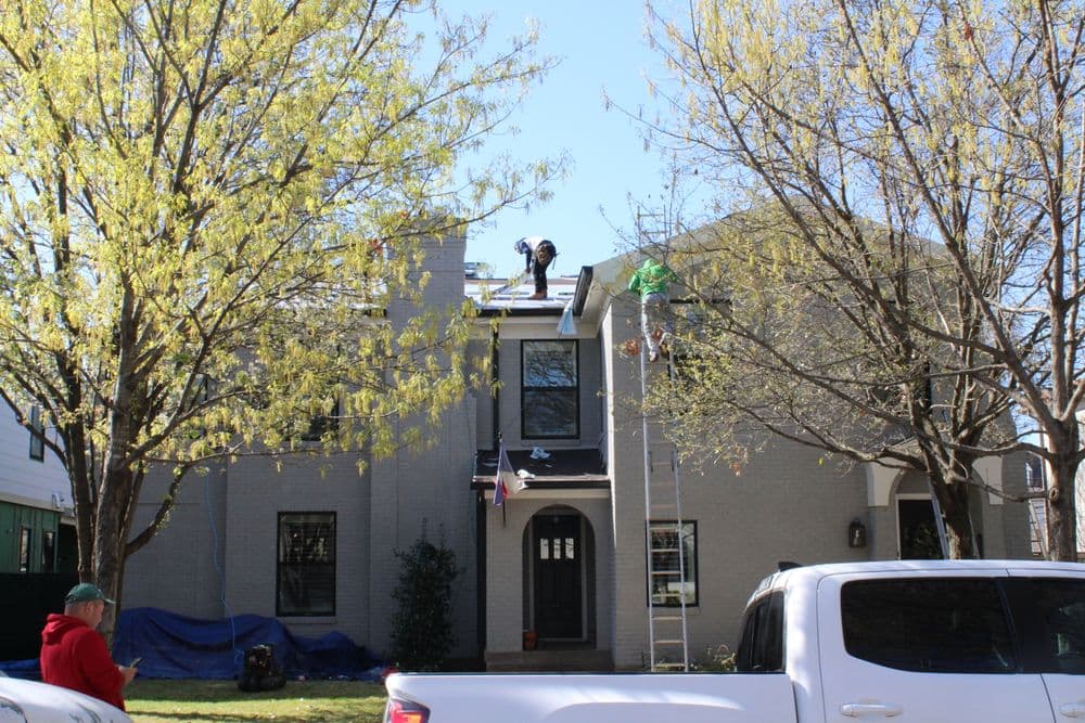Roofing contractors working on a house with trees and a ladder in the foreground.